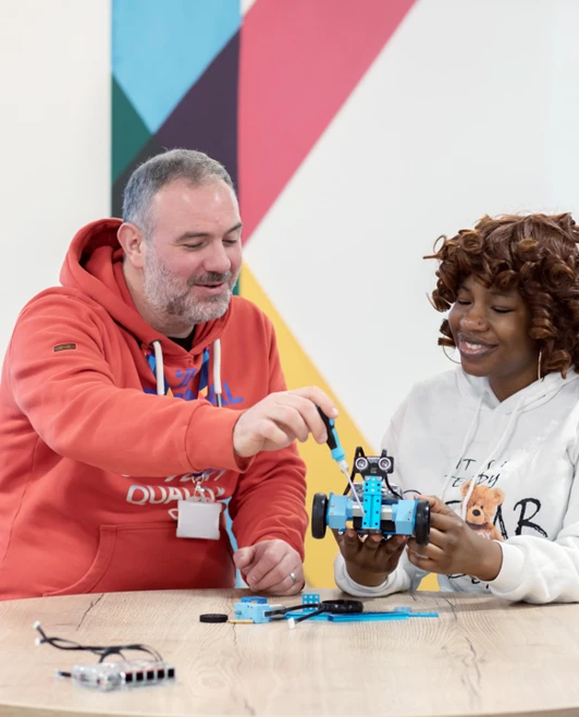 Lecturer in an orange hoodie guiding a student in a white hoodie as they work on a small blue robotic vehicle together. Lecturer in an orange hoodie guiding a student in a white hoodie as they work on a small blue robotic vehicle together.